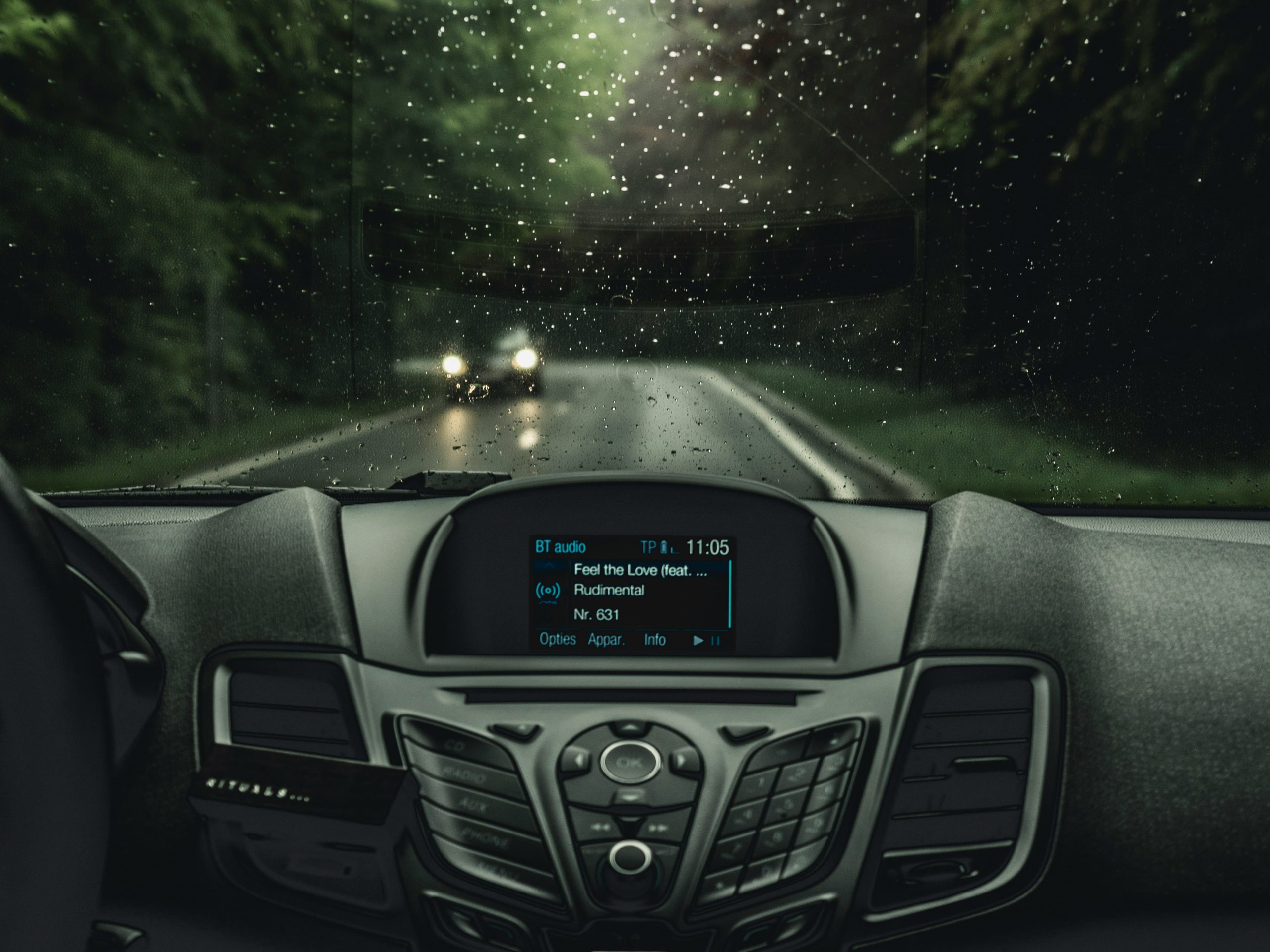 Car interior at night showing a head unit display playing music, with a wet road visible through the windscreen