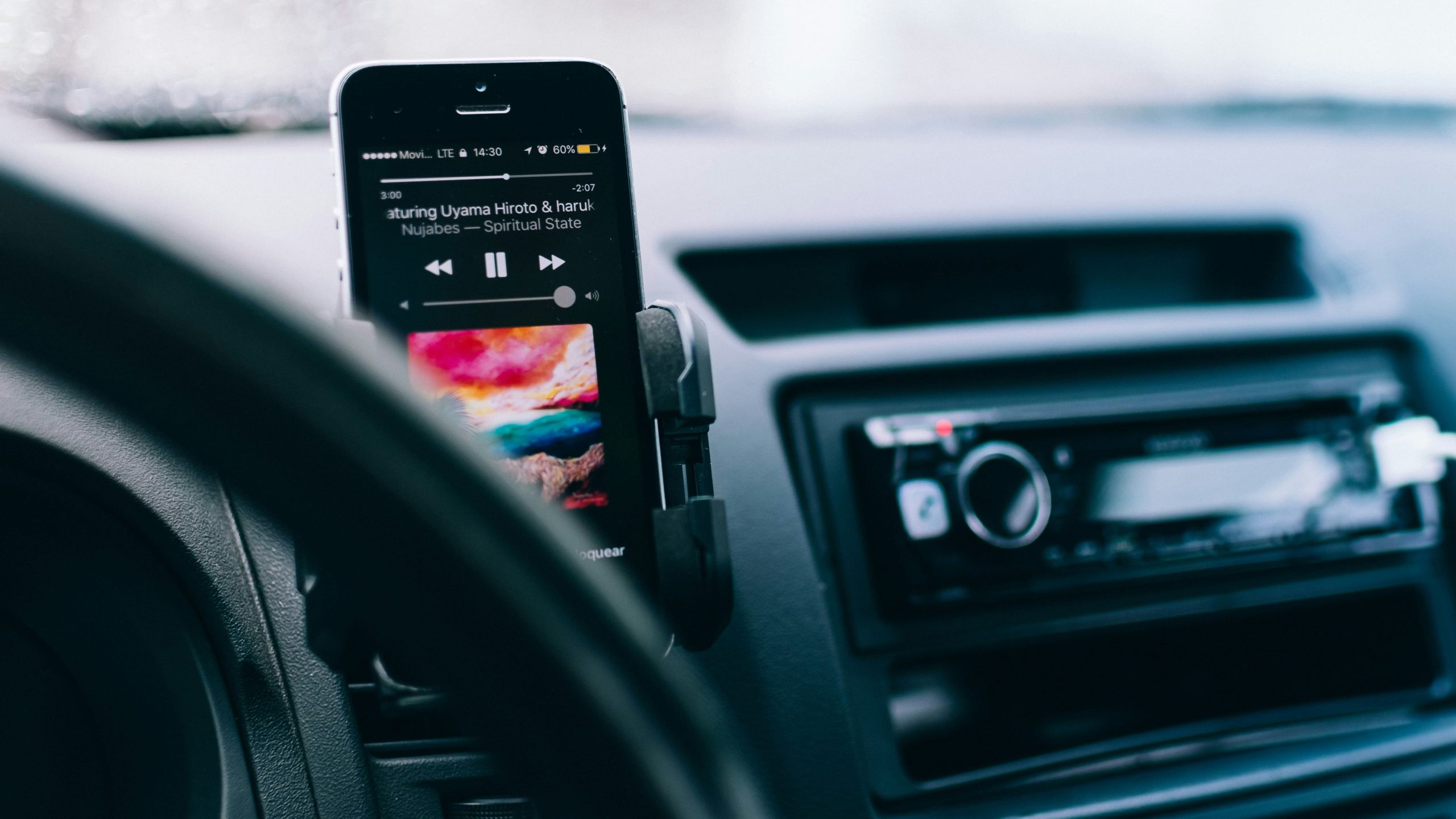 A smartphone mounted near a steering wheel displaying a music player app, serving as the audio source for a traditional aftermarket car audio system receiver in the background.