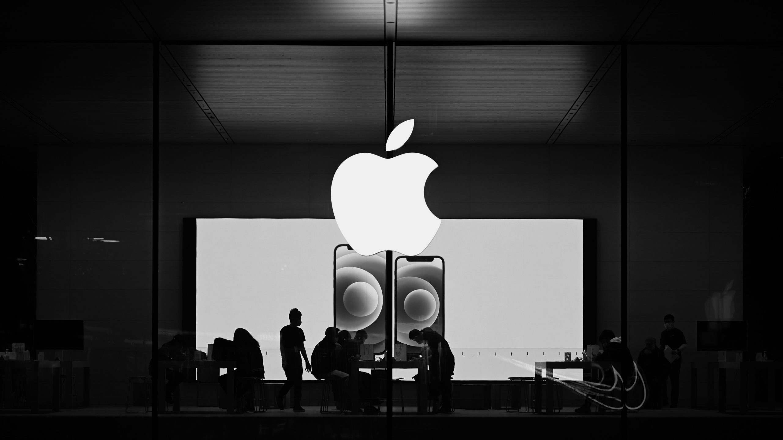 A black and white photograph of an Apple Store exterior, representing the ecosystem powering Apple CarPlay. A large, glowing Apple logo is prominently displayed on the glass window, with silhouettes of customers and staff interacting at tables inside the brightly lit store
